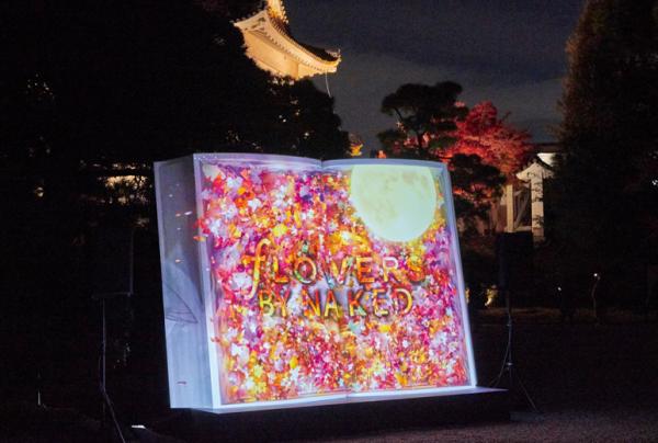 写真：東大手門にある大きな本のオブジェ。色鮮やかな花の背景の上に映し出される「FLOWERS BY NAKED」の文字。